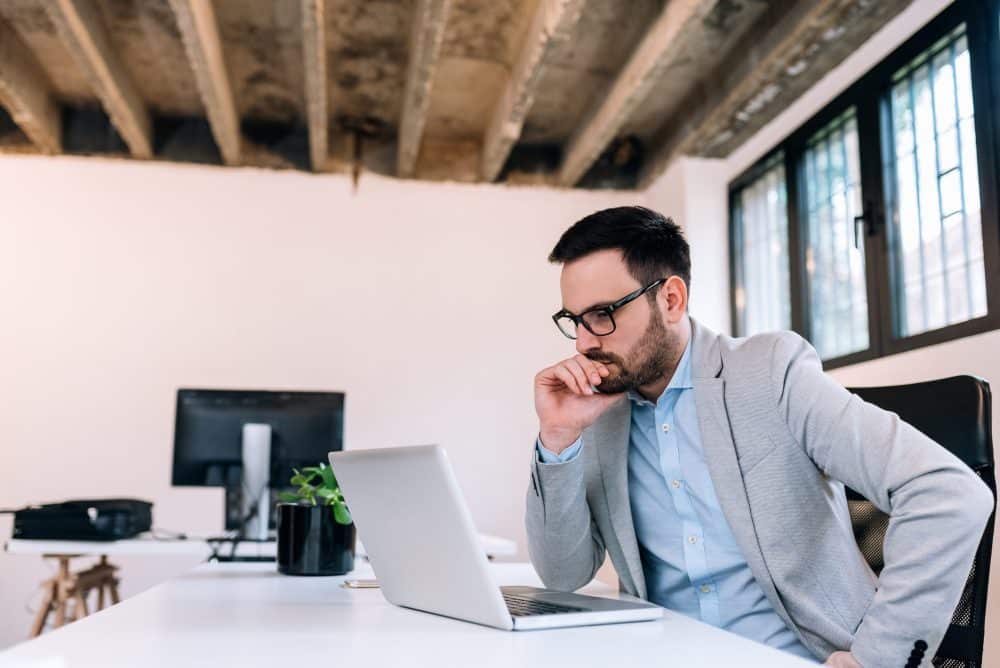 A businessman sitting at a desk and looking thoughtfully at his laptop. 