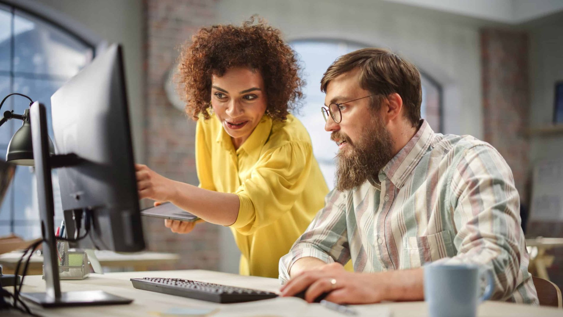 A female employee reviews ROI stats with a male colleague on a desktop computer.